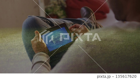 Seated man wearing grey sweater reviewing blueprint on tablet in living room, with red cushion 135617620