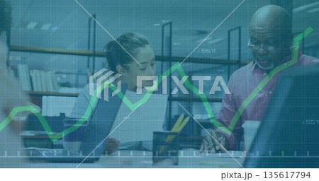 Two coworkers reviewing financial data, working on laptops in office, with documents and coffee cup 135617794