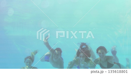 Smiling friends swimming underwater in pool, showing rising bubbles and rippling light patterns 135618801