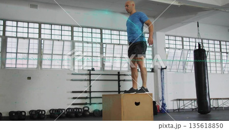 Balancing coach wearing sportswear performing box jump on wooden box in gym, with kettlebells 135618850