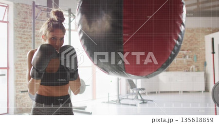 Woman boxer wearing sports bra throwing punches at punching bag in gym studio, with boxing gloves 135618859