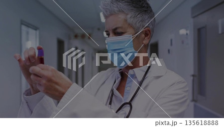 Senior doctor wearing mask inspecting blood sample vial in hospital corridor, with stethoscope Senior doctor wearing mask inspecting blood sample vial in hospital corridor, with stethoscope 135618888