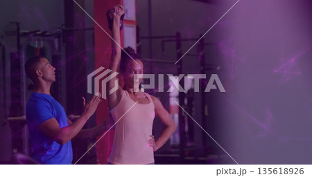 Woman lifting kettlebell overhead in gym area with trainer and squat racks barbells weight plates Woman lifting kettlebell overhead in gym area with trainer and squat racks barbells weight plates 135618926