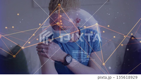 Man wearing blue t-shirt and medical face mask holding left shoulder on couch, with network overlay Man wearing blue t-shirt and medical face mask holding left shoulder on couch, with network overlay 135618929