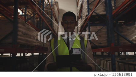 Warehouse inspector scanning pallets on metal racks in narrow aisle, with tablet and safety vest Warehouse inspector scanning pallets on metal racks in narrow aisle, with tablet and safety vest 135618991