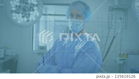 Crossing arms surgeon wearing blue scrubs mask in operating room, with surgical lights and IV stand 135619126
