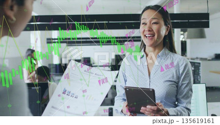Smiling Korean woman holding tablet and discussing financial charts in office, with monitors 135619161