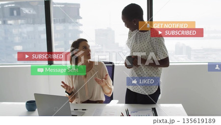Discussing colleagues in business attire gesturing at desk in office, with laptop coffee mug charts Discussing colleagues in business attire gesturing at desk in office, with laptop coffee mug charts 135619189