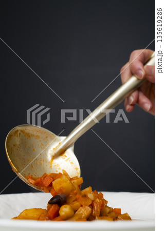 白モツのトマト煮込みをお玉で皿に盛り付ける 135619286