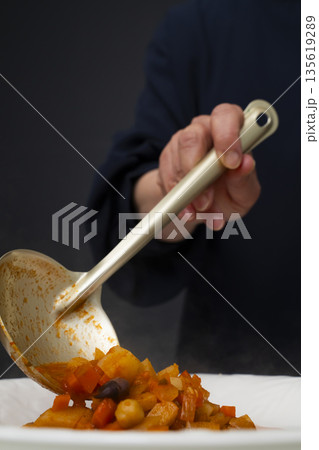 白モツのトマト煮込みをお玉で皿に盛り付ける 135619289