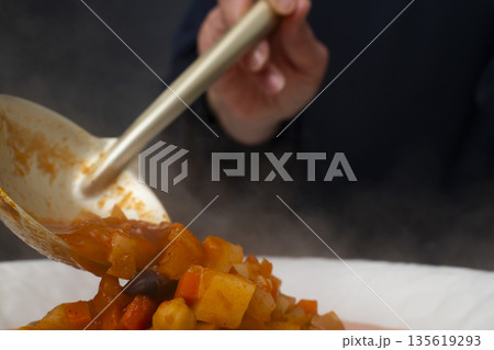 白モツのトマト煮込みをお玉で皿に盛り付ける 135619293