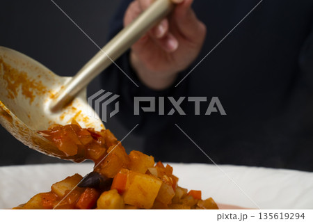 白モツのトマト煮込みをお玉で皿に盛り付ける 135619294