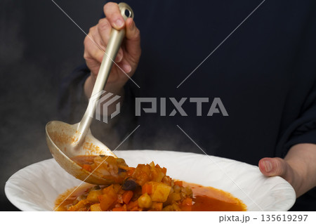白モツのトマト煮込みをお玉で皿に盛り付ける 135619297