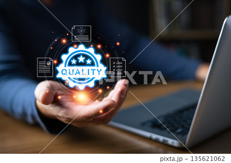 A person working on a laptop presents a glowing digital badge with the word quality surrounded by icons of approved documents, symbolizing excellence, certification, trust, and professional standards. 135621062