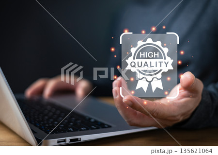 A person using a laptop presents a glowing digital badge labeled high quality, symbolizing trust, excellence, standards, certification, and assurance in business, products, and professional services. 135621064