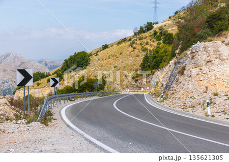 A winding mountain road bends through rocky hills with guardrails and chevron signs 135621305