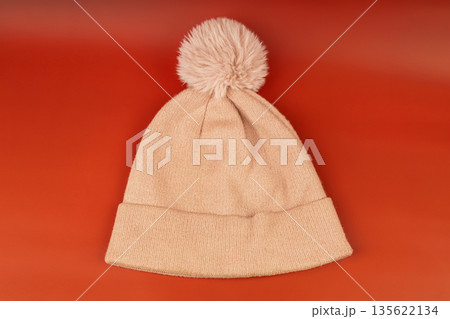 Pink knitted winter hat with a pompom on a red background. Warm wool beanie for cold weather. 135622134
