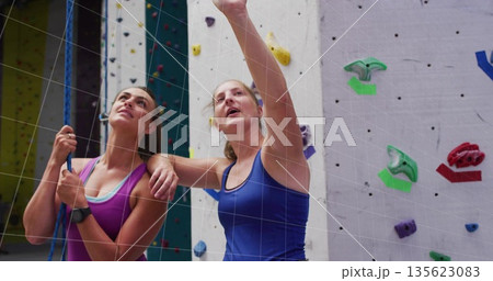 Pointing instructor guiding belaying climber at climbing gym wall, with rope harness and handholds 135623083
