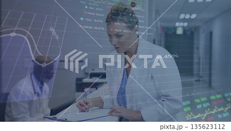 Woman in white lab coat writing on clipboard at nursing station, with stethoscope, data charts 135623112