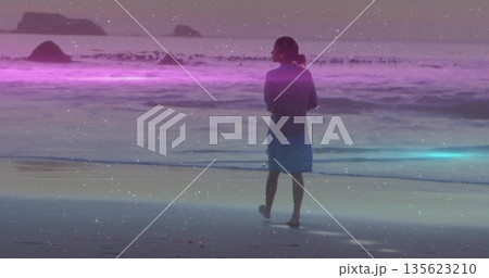 Standing barefoot woman in blue dress facing sea on beach at dusk, purple-blue band and rocks 135623210