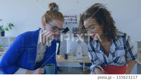 Leaning over desk two female coworkers in blazers examining code in office, with ceramic coffee mug 135623217
