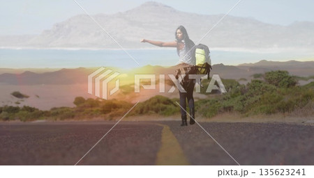 Hitchhiking female traveler extending thumb on paved coastal road, with backpack and plaid shirt Hitchhiking female traveler extending thumb on paved coastal road, with backpack and plaid shirt 135623241