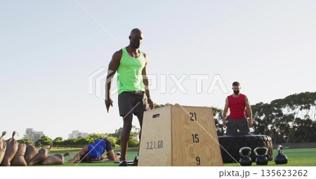 Working out man in green tank top beside plyometric box on park grass, with fitness equipment Working out man in green tank top beside plyometric box on park grass, with fitness equipment 135623262