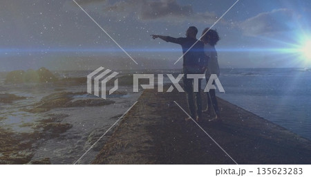 Pointing man and leaning woman gazing across rocky shoreline water at pier, lens flare from sunset 135623283