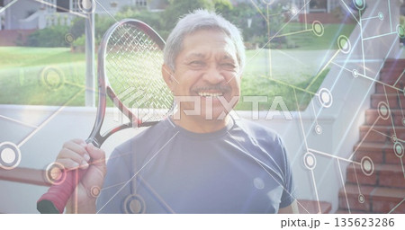 Smiling senior Hispanic man holding tennis racket over shoulder on backyard patio, network overlay Smiling senior Hispanic man holding tennis racket over shoulder on backyard patio, network overlay 135623286