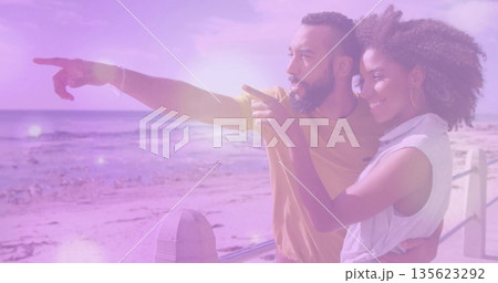 Pointing couple wearing white top and mustard tee on boardwalk featuring railing and ocean horizon 135623292