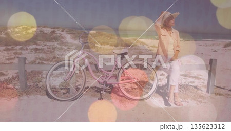 Woman adjusting straw sun hat at dune path, with pink cruiser bicycle and fence, copy space 135623312