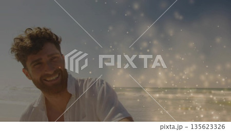 Smiling bearded man wearing white shirt standing on shoreline, sparkling light overlay, copy space 135623326