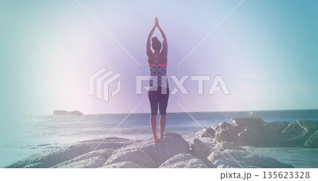 Standing woman stretching in workout top, leggings on rocky shoreline, with ocean horizon backdrop 135623328
