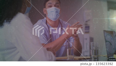 Speaking doctor in scrubs and face mask at clinic desk, with stethoscope laptop papers water 135623392