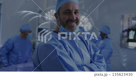 Standing lead surgeon in blue scrubs folding arms in operating room, with overhead lights, monitors Standing lead surgeon in blue scrubs folding arms in operating room, with overhead lights, monitors 135623430