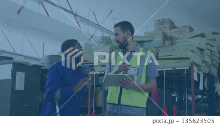 Inspecting warehouse worker in jumpsuit leaning on crutch, supervisor in safety vest holding tablet 135623505