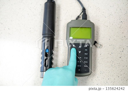 Female hands in protective gloves adjusting settings on a handheld pH meter. 135624242
