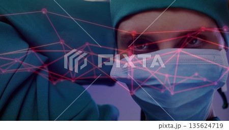 Surgeon in green scrubs, cap adjusting blue glove, mask in operating room with pink network overlay Surgeon in green scrubs, cap adjusting blue glove, mask in operating room with pink network overlay 135624719