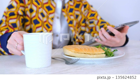 Holding white mug scrolling tablet wearing printed jacket at cafe table with panini fork and greens Holding white mug scrolling tablet wearing printed jacket at cafe table with panini fork and greens 135624852
