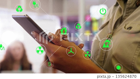 Businesswoman wearing blazer tapping tablet in office, with floating sustainable energy icons 135624870
