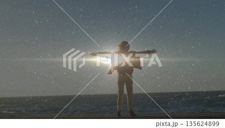 Stretching lone figure wearing jacket extending arms on sandy beach, with sun flare and ocean 135624899