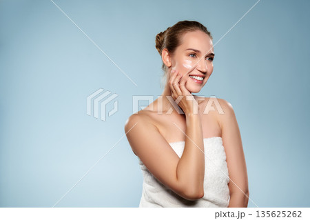 Woman smiling while applying skincare product in a simple setting with light blue background Woman smiling while applying skincare product in a simple setting with light blue background 135625262