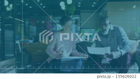 Two seated colleagues reviewing printed documents on sofa in office lounge, holding coffee cup 135626022