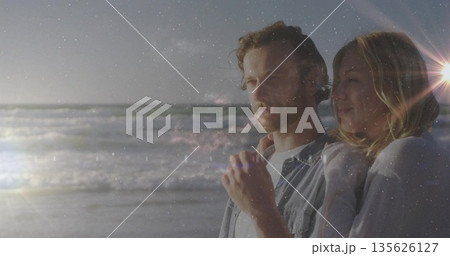 Gazing denim-clad man and sweater-clad woman on beach with waves sinking sun lens flare, copy space 135626127