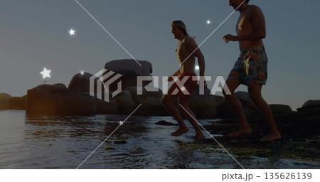 Stepping two friends in swim trunks exploring tide pools at water's edge, with boulders and seaweed 135626139
