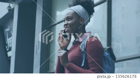 Talking woman wearing burgundy sweater holding smartphone on urban sidewalk, carrying blue backpack 135626178