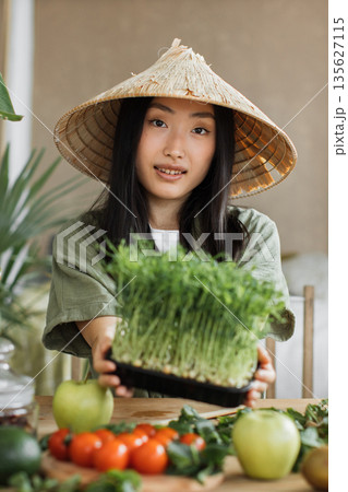 A young Asian woman wearing a conical hat holds a tray of fresh microgreens, surrounded by fruits and vegetables on a wooden table A young Asian woman wearing a conical hat holds a tray of fresh microgreens, surrounded by fruits and vegetables on a wooden table 135627115