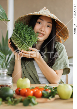 A young Asian woman wearing a conical hat holds a tray of fresh microgreens while surrounded by healthy vegetables and fruits on a wooden table A young Asian woman wearing a conical hat holds a tray of fresh microgreens while surrounded by healthy vegetables and fruits on a wooden table 135627116