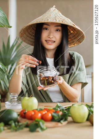 A young Asian woman wearing a traditional conical hat holds a jar of dates and prepares fresh produce for a healthy meal A young Asian woman wearing a traditional conical hat holds a jar of dates and prepares fresh produce for a healthy meal 135627120