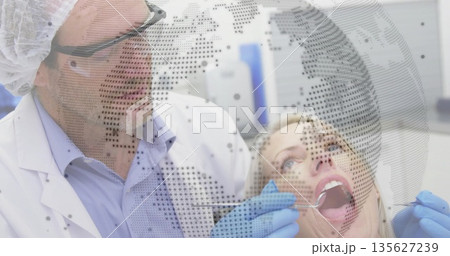 Examining male dentist leaning over female patient in dental clinic, with mirror and globe overlay Examining male dentist leaning over female patient in dental clinic, with mirror and globe overlay 135627239